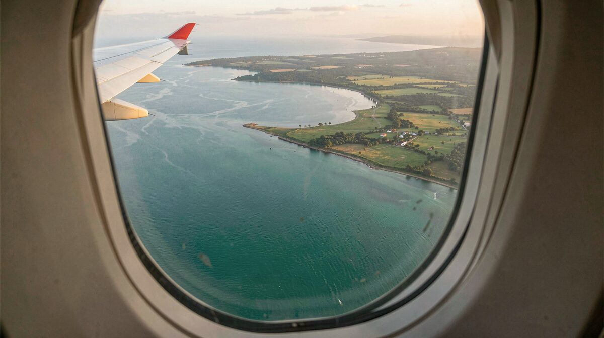 Vue aérienne depuis un hublot d’avion d’une côte ouest-africaine, mer turquoise vertes rencontrant des terres agricoles et l’embouchure d’un large fleuve, aile blanche à bout rouge visible